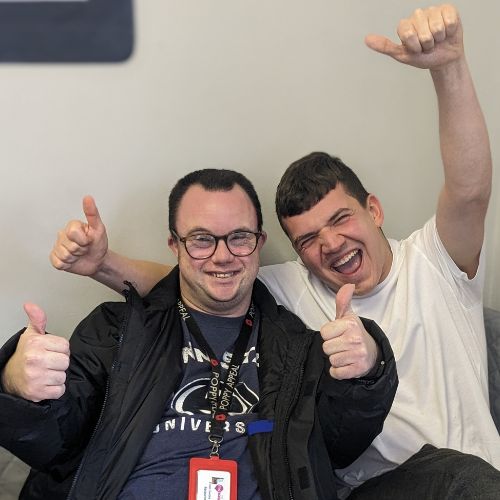Two men smiling with their thumbs up sitting on a sofa