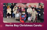 A group of members from East Kent Mencap’s Herne Bay hub stand together outdoors, many wearing festive Christmas hats and smiling.