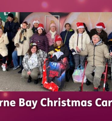 A group of members from East Kent Mencap’s Herne Bay hub stand together outdoors, many wearing festive Christmas hats and smiling.